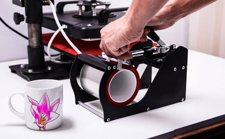 Eine Becherpresse in Schwarz mit rotem Element, während eine Hand den Griff betätigt; daneben eine weiße Tasse mit pinker Orchideenblüte