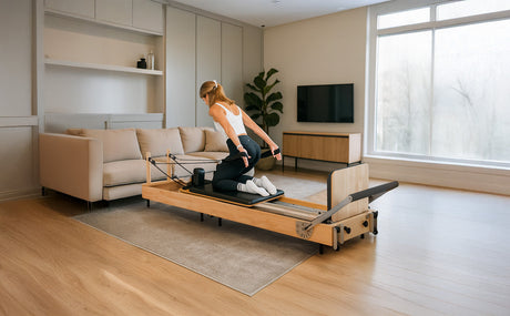 Moderne Pilates Reformer Maschine aus hellem Holz, mit einer sportlichen Frau in Schwarz, die eine Dehnübung im stilvollen Wohnzimmer ausführt
