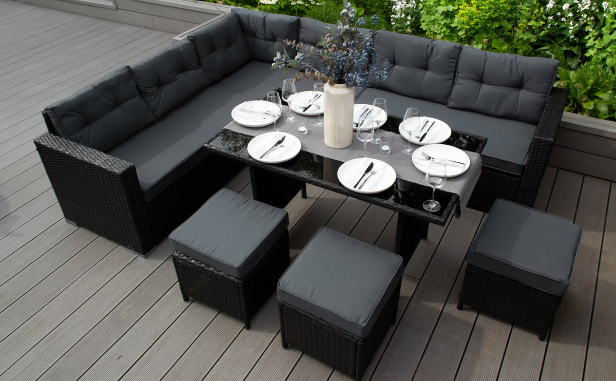 Elegantes Rattanmöbel-Set mit grauen Kissen, schwarzem Tisch und Stühlen, umgeben von grünen Pflanzen auf einer Holzterrasse