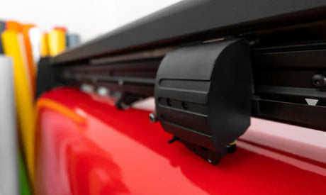 Nahaufnahme eines Vinylschneiders mit schwarzem Gehäuse und rotem Untergrund, umgeben von bunten Vinylrollen für personalisierte Produkte