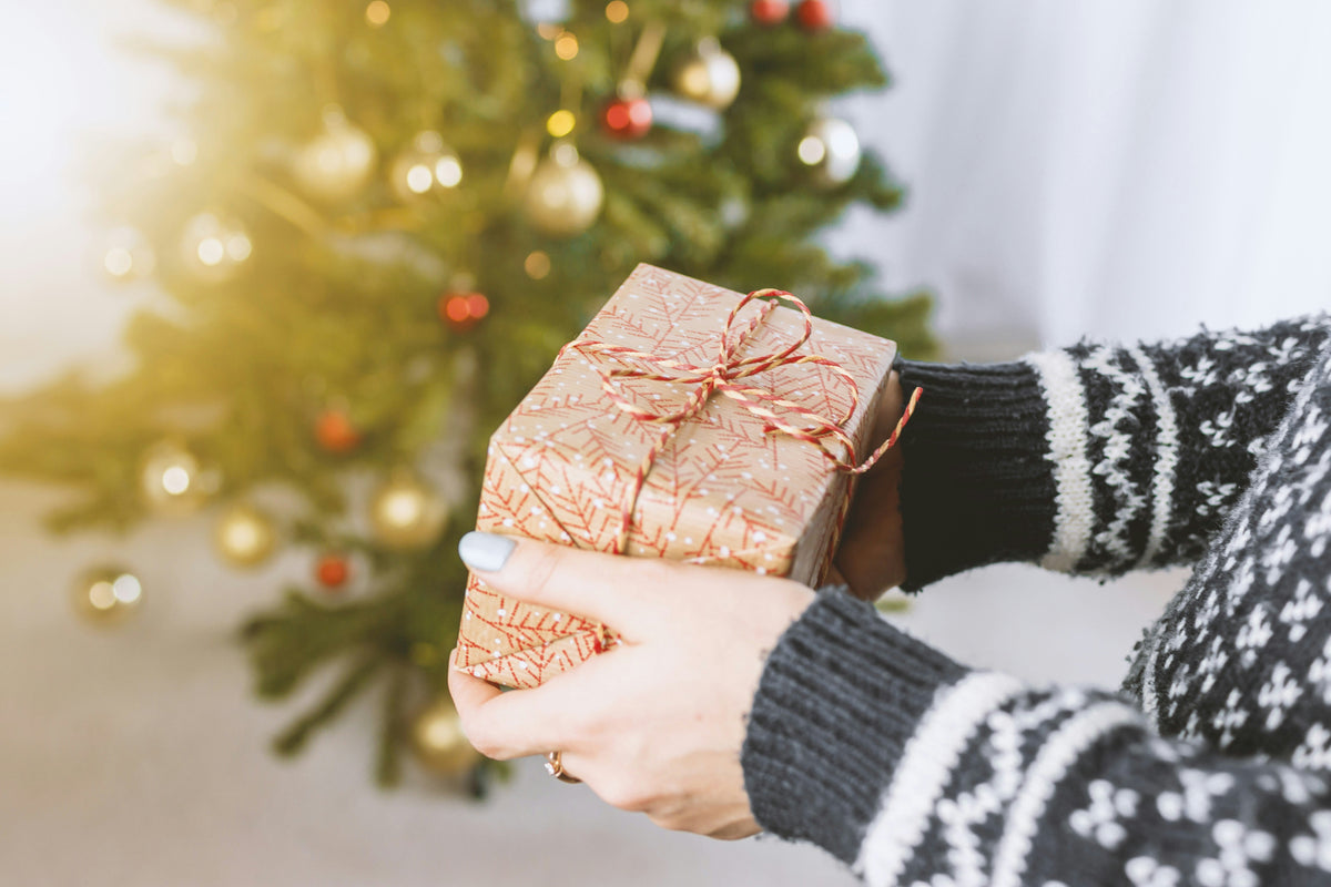 Eine Person hält ein festlich verpacktes Geschenk mit goldenem Papier und rotem Band vor einem Weihnachtsbaum mit bunten Ornamenten