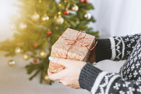 Eine Person hält ein festlich verpacktes Geschenk mit goldenem Papier und rotem Band vor einem Weihnachtsbaum mit bunten Ornamenten
