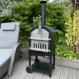 Schwarzer KuKoo Outdoor Pizzaofen mit silberner Front, robustem Design, auf Holzdeck neben einer Liege, umgeben von grünen Pflanzen und Blumen