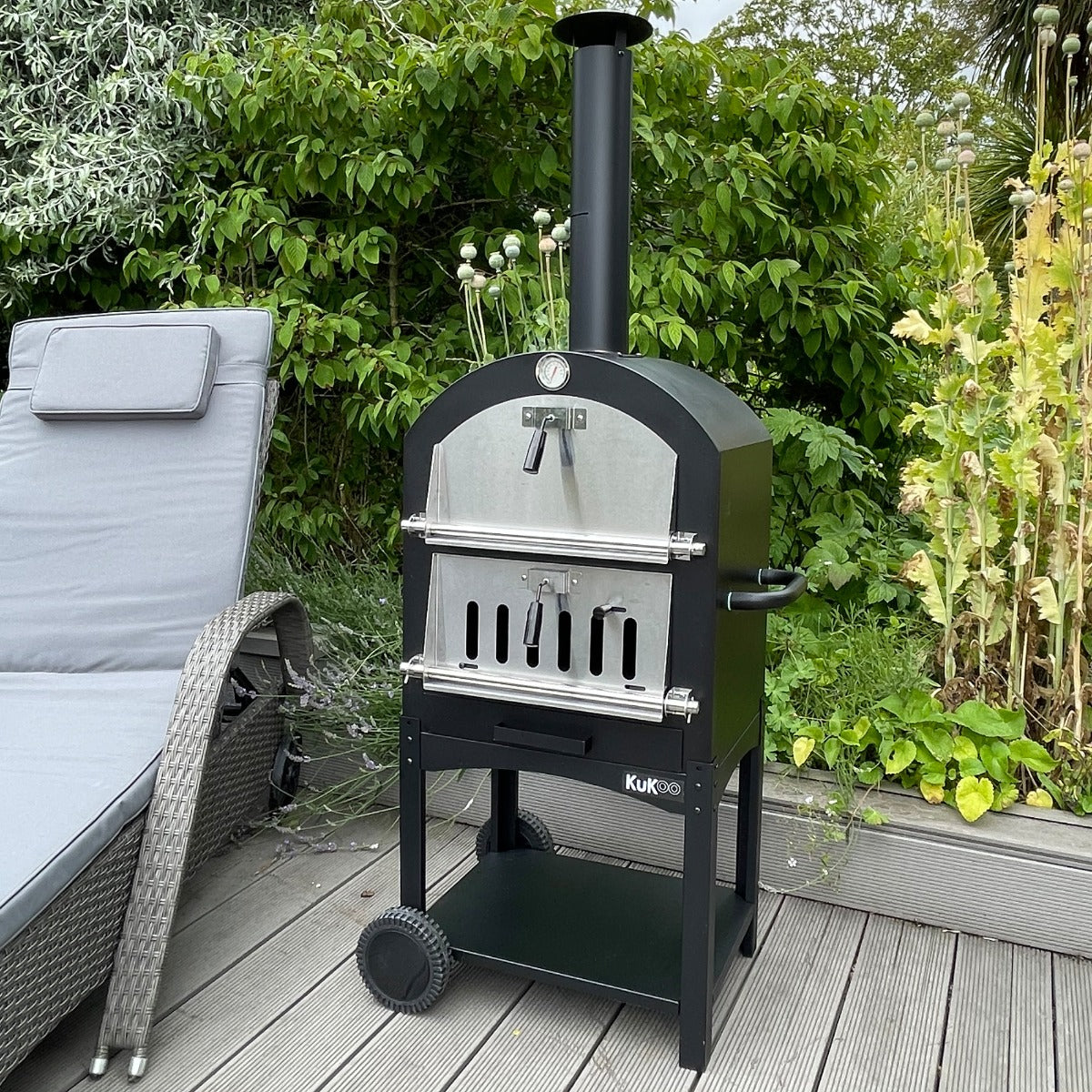 Schwarzer KuKoo Outdoor Pizzaofen mit Edelstahlfront, auf einem Holzdeck platziert, umgeben von grünen Pflanzen und einem gemütlichen Liegestuhl