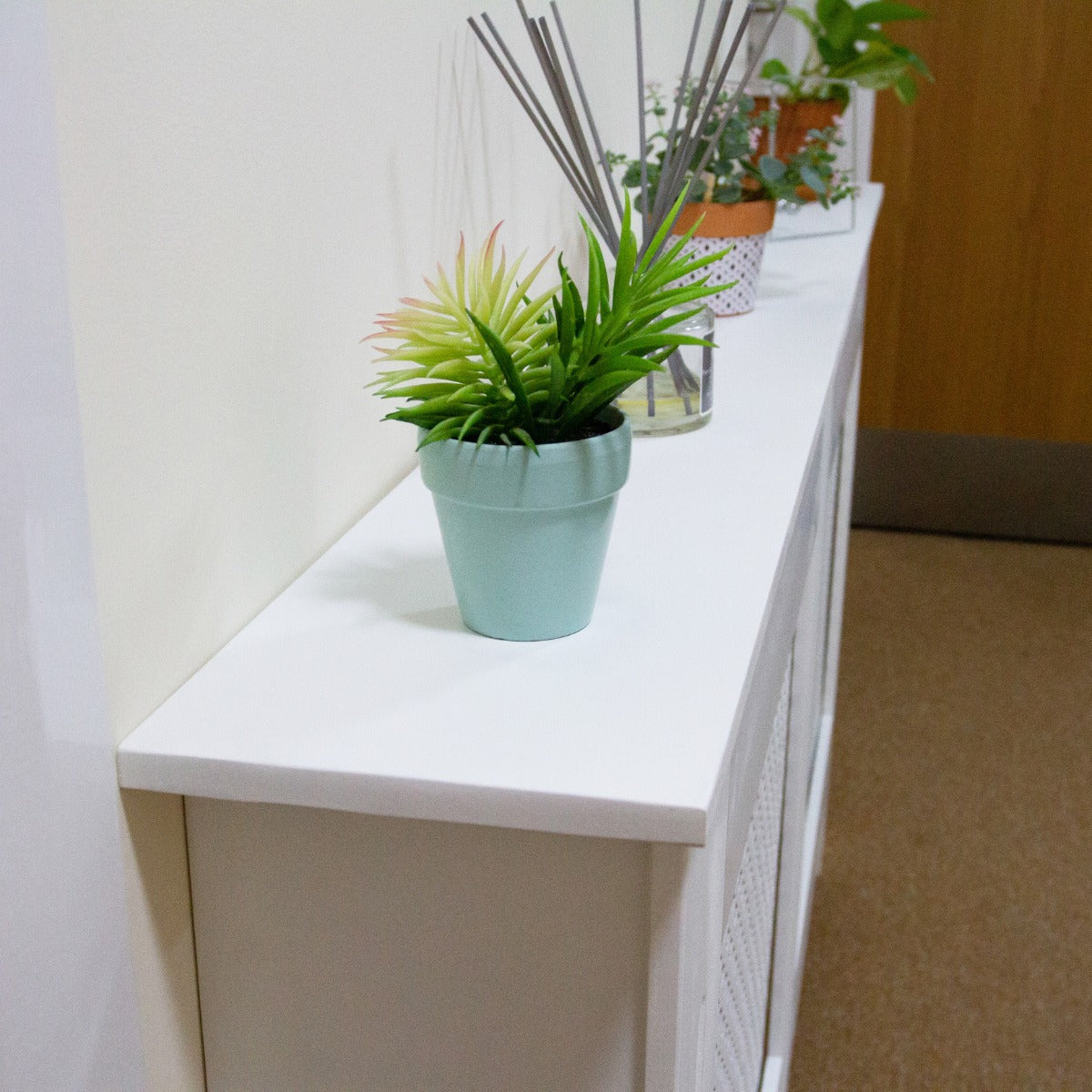 Weiße Heizkörperverkleidung aus Holz mit glatter Oberfläche, dekoriert mit einem hellblauen Blumentopf und einer grünen Pflanze, ideal für moderne Räume
