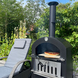 Schwarzer KuKoo Outdoor Pizzaofen mit Holzfeuer, auf dem eine frische Pizza backt, umgeben von grünen Pflanzen und einem gemütlichen Stuhl
