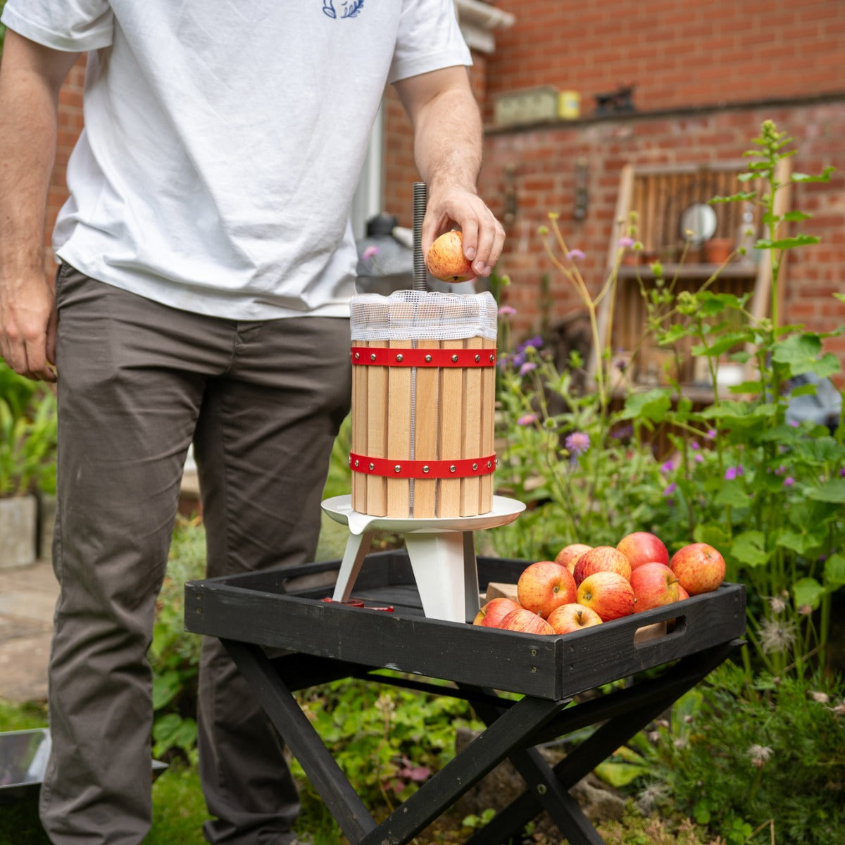 Holzobstpresse mit rotem Band, auf einem weißen Ständer, während ein Apfel hineingelegt wird; im Hintergrund ein Garten mit Äpfeln auf einem Tisch