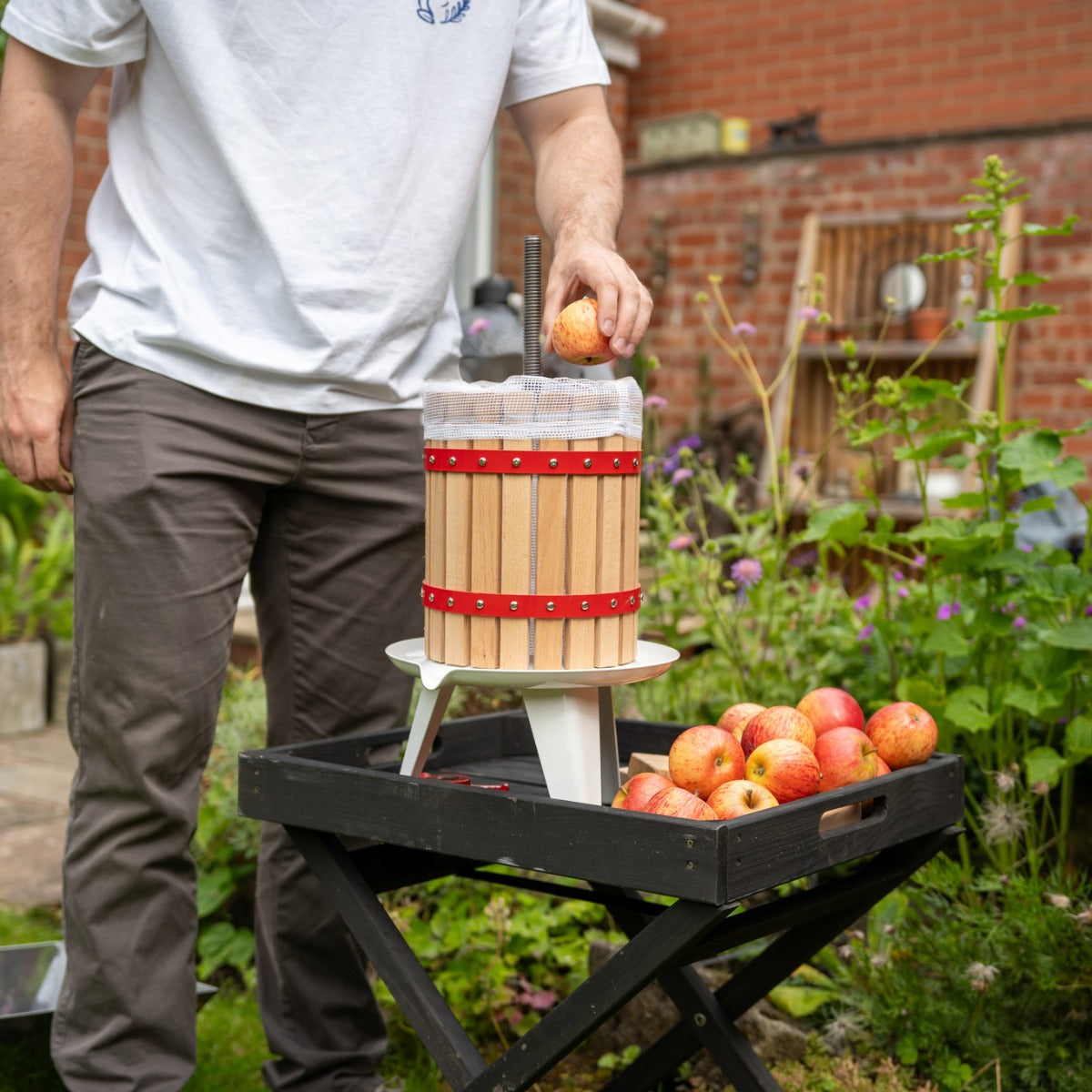 Holz-Obstpresse mit rotem Band, auf einem Tisch im Garten, während eine Person einen Apfel hineinlegt, um frischen Saft zu pressen