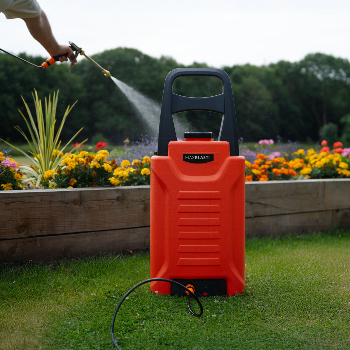 Der orangefarbene 30L Wasserwagen mit ergonomischem Griff steht auf grünem Rasen, umgeben von bunten Blumen, während Wasser gespritzt wird