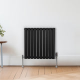 Schwarzer ovaler Röhrenheizkörper mit Chrom-Thermostatventilen, 600mm x 600mm, an einer hellen Wand, umgeben von einem Holzfußboden und einer Pflanze