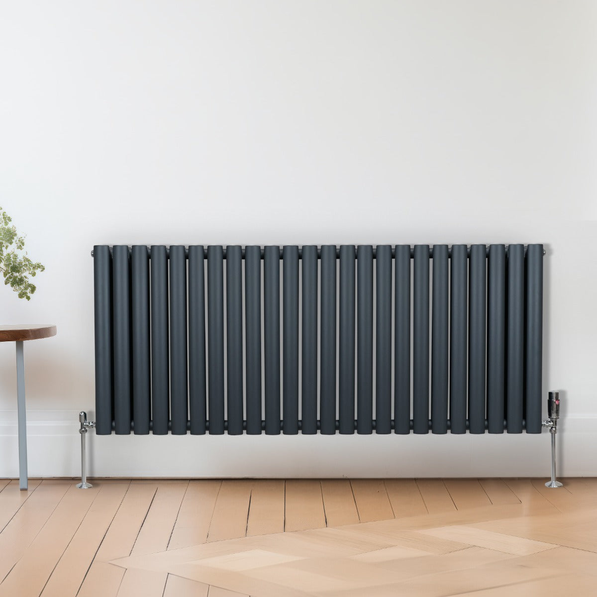 Ovaler Röhrenheizkörper in Anthrazitgrau mit Chrom-Thermostatventilen, 600mm x 1440mm, an einer hellen Wand, auf einem Holzfußboden