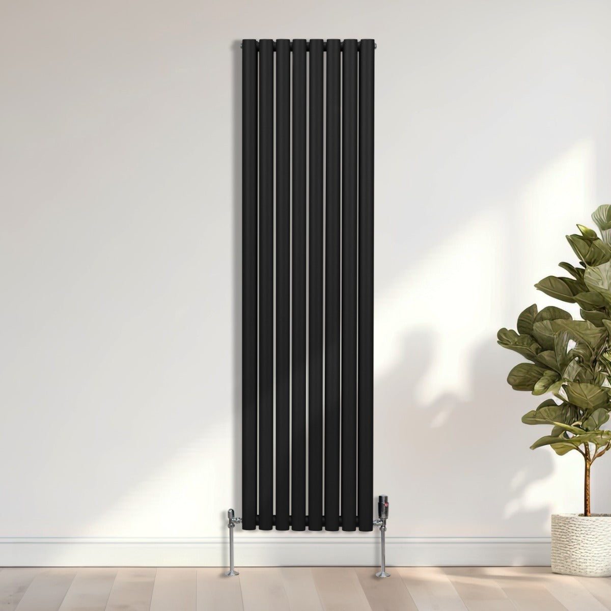 Schwarzer ovaler Röhrenheizkörper mit Chrom-Thermostatventilen, 1800mm x 480mm, vertikal an einer hellen Wand, umgeben von Pflanzen