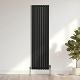 Schwarzer ovaler Röhrenheizkörper mit Chrom-Thermostatventilen, 1800mm x 480mm, vertikal an einer hellen Wand, umgeben von Pflanzen
