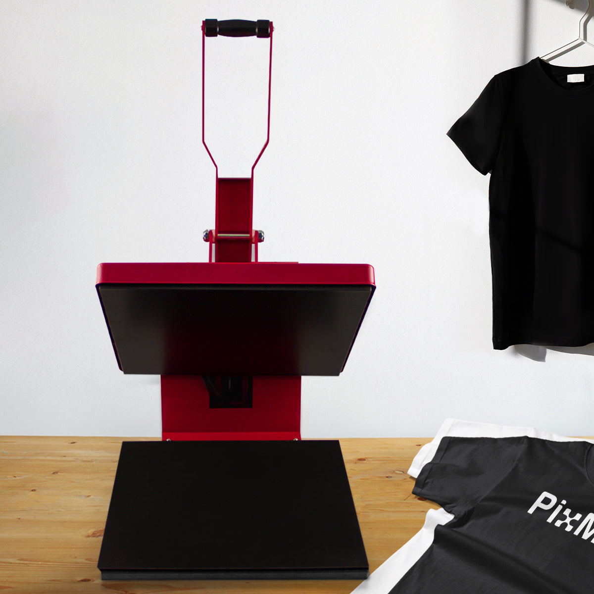 Rote Transferpresse mit schwarzer Heizplatte, ideal für T-Shirt-Druck, auf einem Holztisch, neben einem grauen und einem schwarzen Shirt