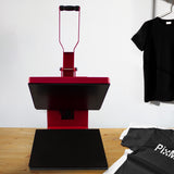 Rote Transferpresse mit schwarzer Heizplatte, ideal für T-Shirt-Druck, auf einem Holztisch, neben einem grauen und einem schwarzen Shirt