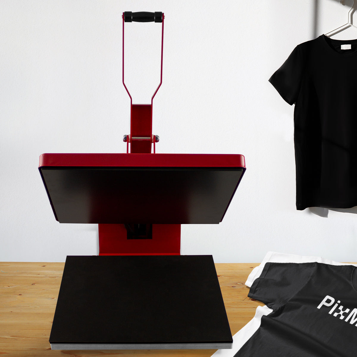 Rote PixMax Transferpresse mit schwarzer Heizplatte, ideal für Textildruck, auf einem Holztisch neben einem schwarzen T-Shirt und einem weißen Shirt