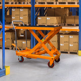 Hydraulischer Hubtisch in leuchtendem Orange, mit stabiler Plattform und Rollen, steht vor Regalen voller Kartons in einem Lagerraum