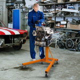 Robuster T-Mech 900kg Motorständer in leuchtendem Orange, unterstützt einen Mechaniker bei der Arbeit an einem Motor in einer Werkstatt mit Werkzeugen und Fahrzeugen im Hintergrund
