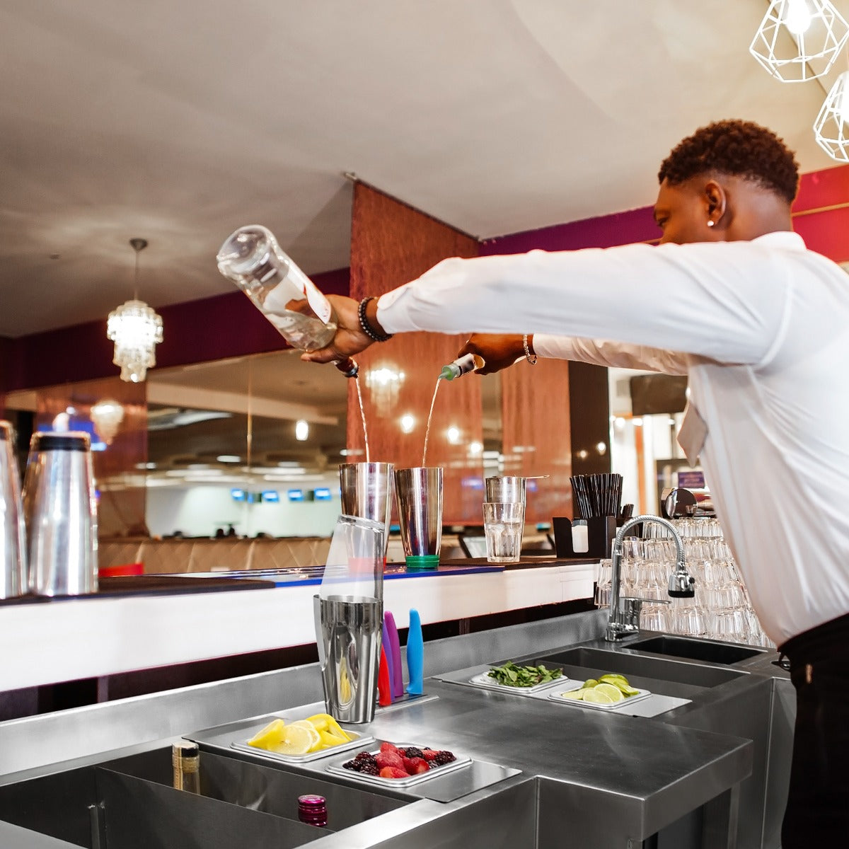 Ein Barkeeper mixt Cocktails an einer modernen, kompakten Duo-Barstation aus Edelstahl, umgeben von frischen Zutaten wie Zitrone, Minze und Beeren