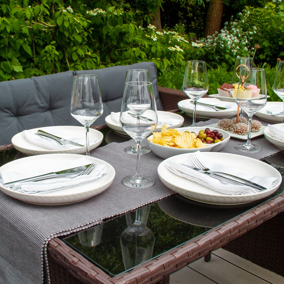 Elegantes Polyrattan Gartenmöbel Set in Braun, gedeckter Tisch mit weißen Tellern, Gläsern und Snacks, umgeben von grüner Natur und Blumen