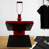 Rote PixMax Transferpresse mit schwarzer Heizplatte, auf einem Holztisch platziert, neben einem grauen T-Shirt und einer PTFE-Matte
