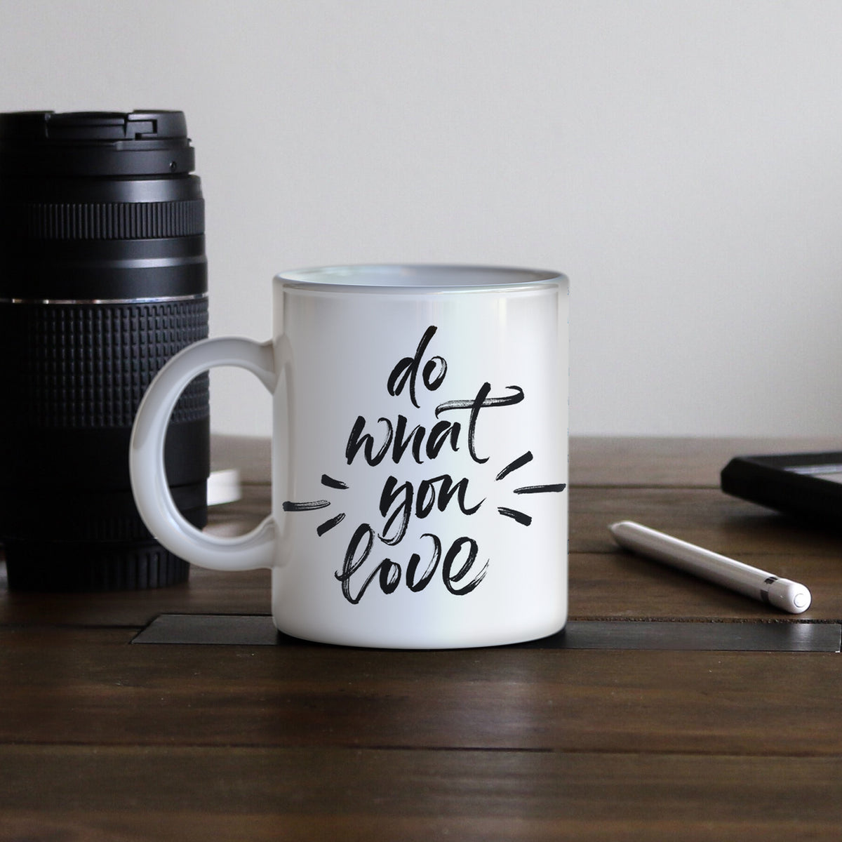 Weißer Becher mit schwarzem Schriftzug "do what you love", auf einem Holztisch neben einer Kamera und einem Notizbuch platziert