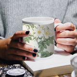 Weißer Becher mit floralen Motiven, gehalten von einer Person mit schwarzen Nägeln, auf einem Holztisch neben einem Buch und einer Tasse