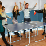 Tischtennisplatte in einem modernen Raum mit fünf Personen, die aktiv spielen, zwei rote Bälle und eine blaue Oberfläche, fröhliche Stimmung
