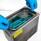 Ultraschallreiniger mit blauer Flüssigkeit, Edelstahlkorb und Händen in blauen Handschuhen, die den Korb herausheben