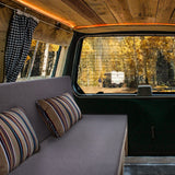 Gemütliches Bett im Camper Van mit grauem Stoffbezug und gestreiften Kissen, umgeben von Holzverkleidung und sanfter Beleuchtung