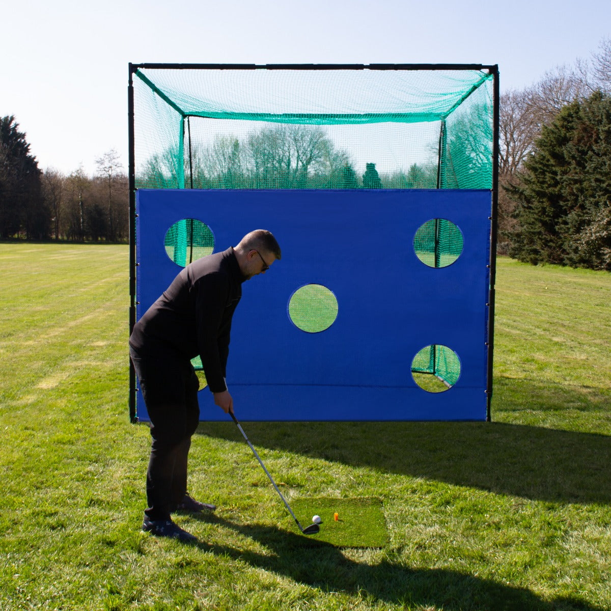 Ein Golfer steht vor einem blauen Golfkäfignetz mit runden Löchern, auf einer grünen Wiese, bereit, seinen Ball zu schlagen