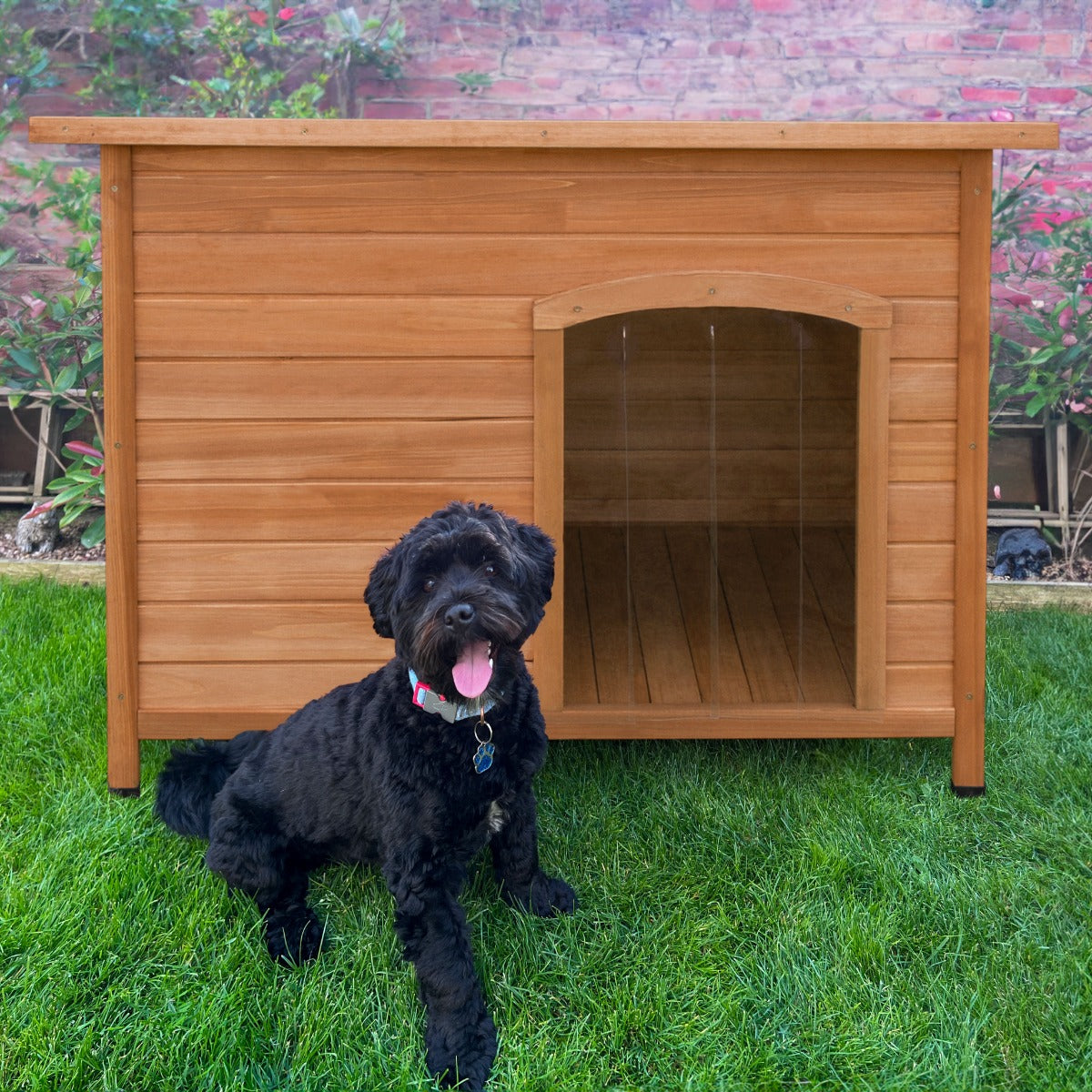 Große Hundehütte aus hellem Holz mit einer offenen Tür, daneben ein schwarzer Hund auf grünem Rasen, umgeben von Pflanzen