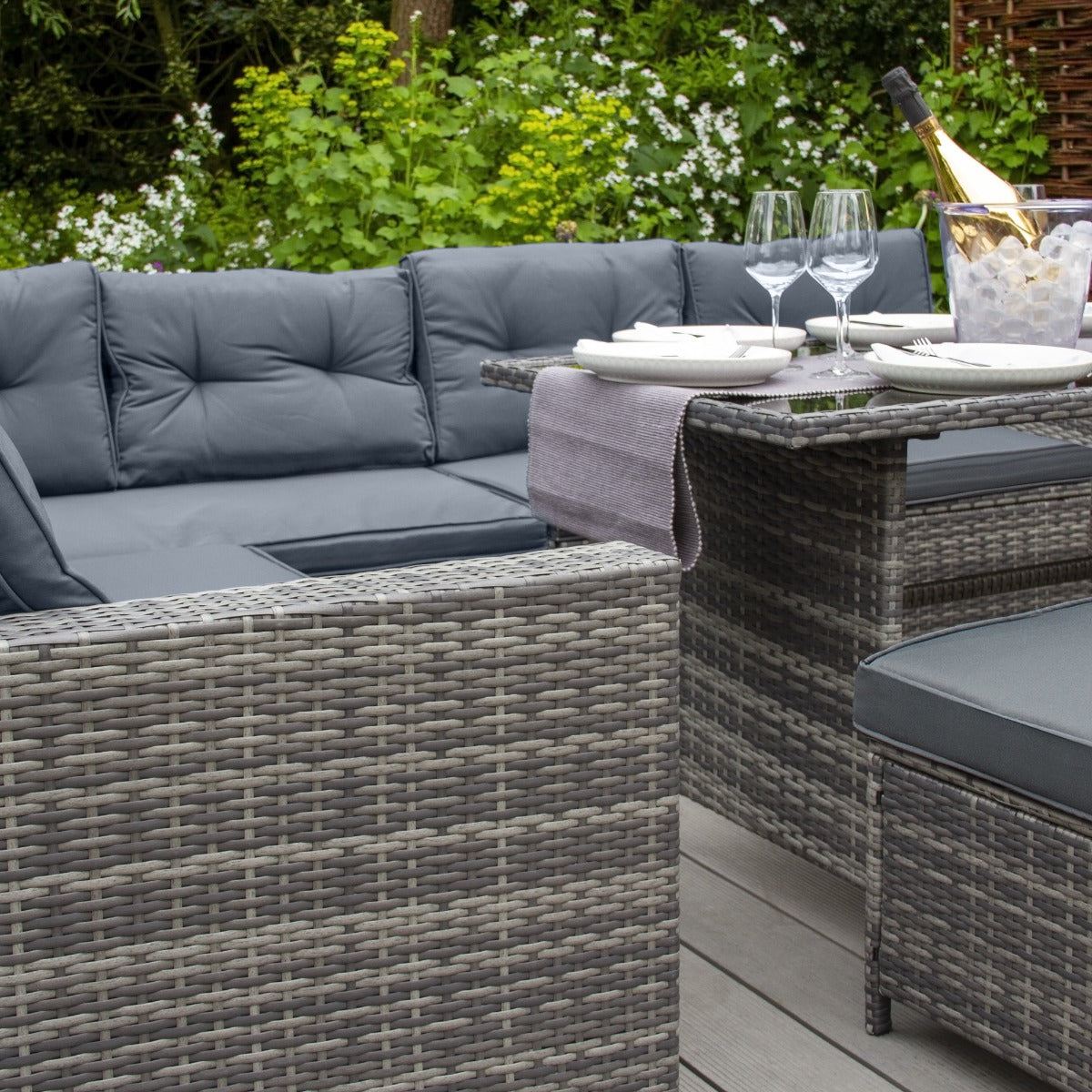 Grau polyrattan Gartenmöbel Set mit bequemen Kissen, eleganter Tisch, Gläsern und einer Flasche Sekt, umgeben von grüner Pflanzenlandschaft