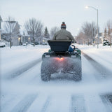 Ein Quad mit einem ATV-Streuer fährt durch eine verschneite Straße, während Schnee auf die Fahrbahn gestreut wird, um die Sicht zu verbessern