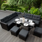 Schwarze Rattan Eckgarnitur mit bequemen, grauen Kissen und einem eleganten Tisch, umgeben von üppigem Grün und blühenden Pflanzen