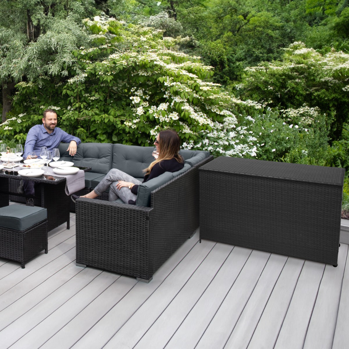 Schwarze Rattan Eckgarnitur mit bequemen Kissen und praktischer Kissenbox, umgeben von grüner Natur auf einer Holzterrasse