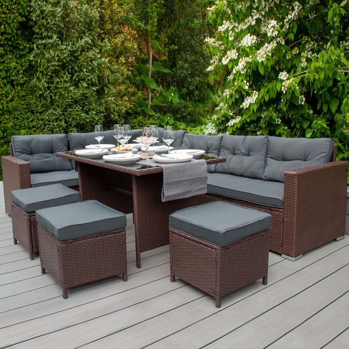 Braune Rattan Eckgarnitur mit bequemen, grauen Kissen und einer großen Tischfläche, umgeben von üppigem Grün im Garten