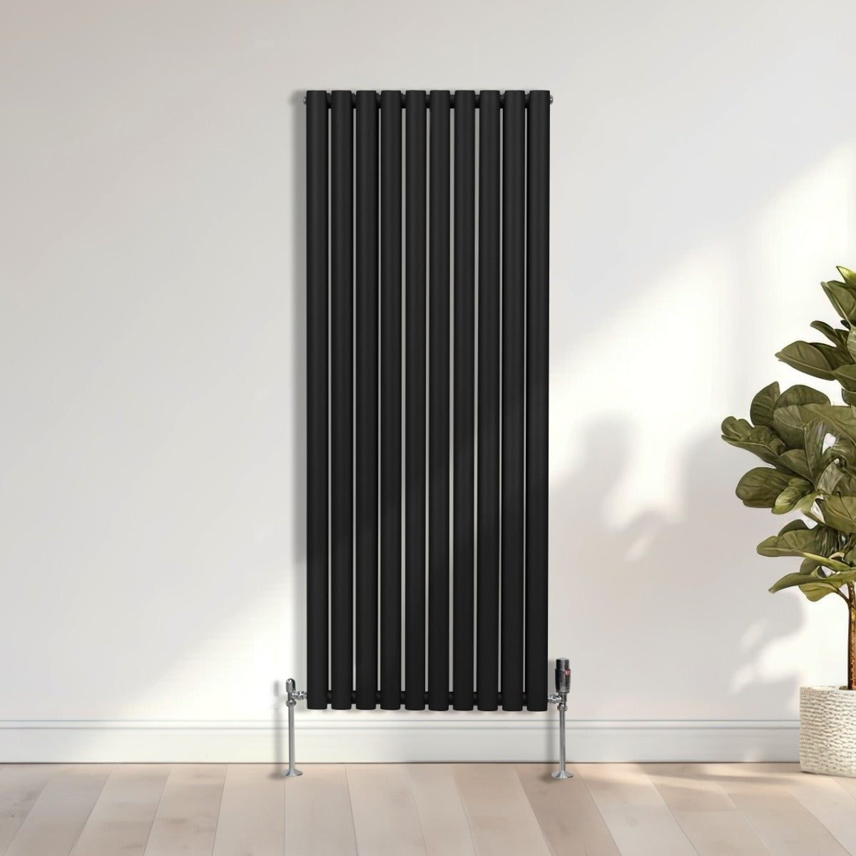 Schwarzer ovaler Röhrenheizkörper mit vertikalen Lamellen, 1600mm x 600mm, elegant an einer hellen Wand montiert, umgeben von einem grünen Pflanzenarrangement
