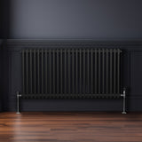 Traditioneller 2-Spalten-Heizkörper in Schwarz, 600 x 1192 mm, mit glänzenden Metallfüßen, vor einer dunklen Wand und auf einem Holzfußboden