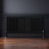 Traditioneller 3-Spalten-Heizkörper in Schwarz, 600 x 1192 mm, mit glänzenden Metallventilen, vor einer dunklen Wand und auf einem Holzfußboden
