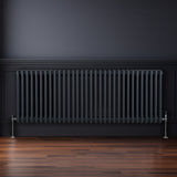 Traditioneller 3-Spalten-Heizkörper in Anthrazitgrau, 600 x 1462 mm, mit glänzenden Metallfüßen, vor einer dunklen Wand und auf einem Holzfußboden