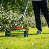 Rasenrakel in leuchtendem Orange mit breiter, rollender Bürste, ideal zum Lüften von Rasenflächen, auf grünem Gras im Hintergrund