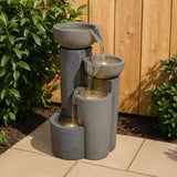 Elegante elektrische Wasseranlage "Tranquil Tiers" mit grauen, strukturierten Behältern und goldenen Wasserströmen, umgeben von grünen Pflanzen