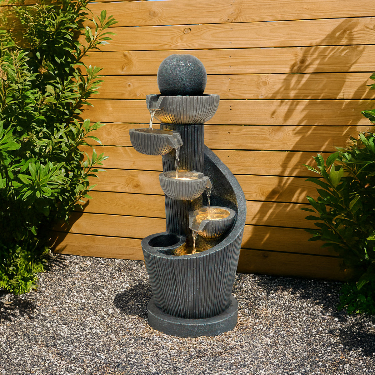 Elegante elektrische Wasseranlage "Spiral Springs" aus schwarzem Stein, mit mehreren Wasserschalen, die sanft über die Stufen fließen, umgeben von grünen Pflanzen
