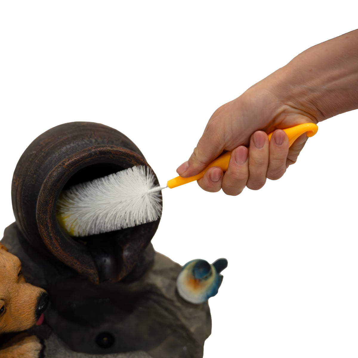 Eine Hand hält eine orangefarbene Bürste, die eine dekorative Wasseranlage mit einem Hund und einem Vogel reinigt, umgeben von Steinen
