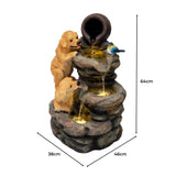 Zwei verspielte Hunde aus Kunststein, die an einer mehrstufigen Wasseranlage mit einem Tontopf und einem blauen Vogel sitzen, in natürlichen Erdtönen gestaltet