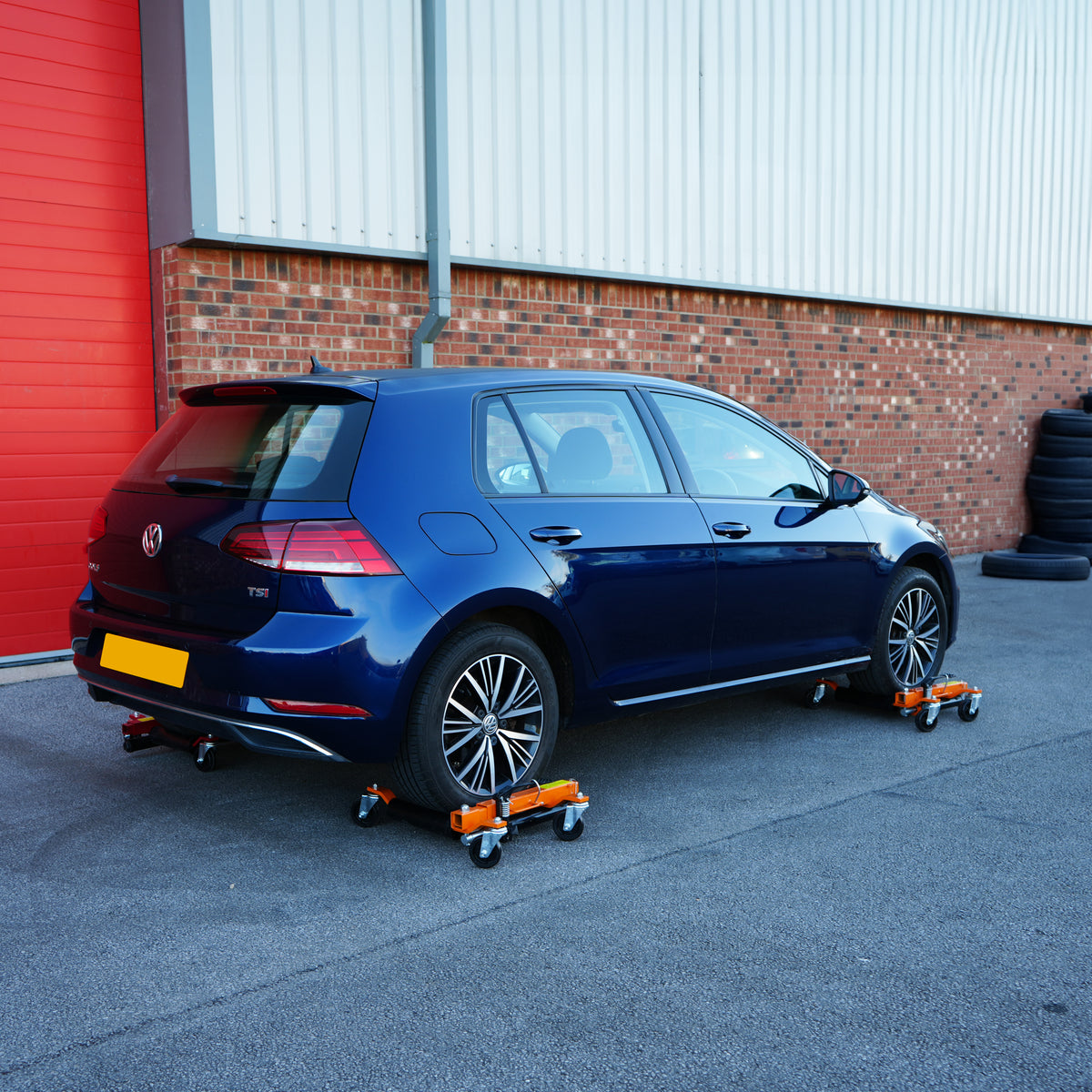 Hydraulische Positionierungsschlitten unter einem blauen Volkswagen Golf, ausgestattet mit stabilen Rollen und orangefarbenen Halterungen auf einem grauen Asphaltboden