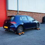 Hydraulische Positionierungsschlitten unter einem blauen Volkswagen Golf, ausgestattet mit stabilen Rollen und orangefarbenen Halterungen auf einem grauen Asphaltboden