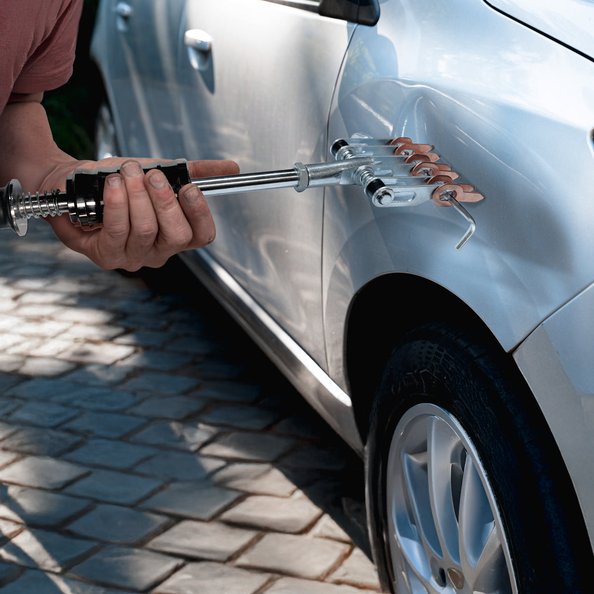 Hand hält einen Ausbeulenzieher mit metallischen Greifern, der auf einer silbernen Autotür angewendet wird, um Dellen zu entfernen