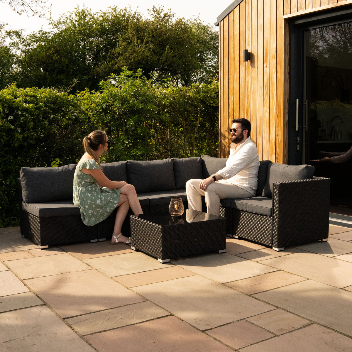 Modulares 7-teiliges Rattan-Möbelset in Schwarz mit grauen Kissen, ideal für den Außenbereich, umgeben von grüner Natur und einem modernen Holzhaus